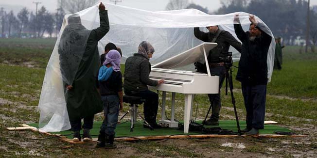 Chinese dissident artist Ai Weiwei (R) holds a rain cover to protect a Syrian refugee woman from the rain, as she performs in a field, on a piano brought by the artist, near a makeshift camp on the Greek-Macedonian border, near the village of Idomeni, Greece March 12, 2016. REUTERS/Stoyan Nenov 