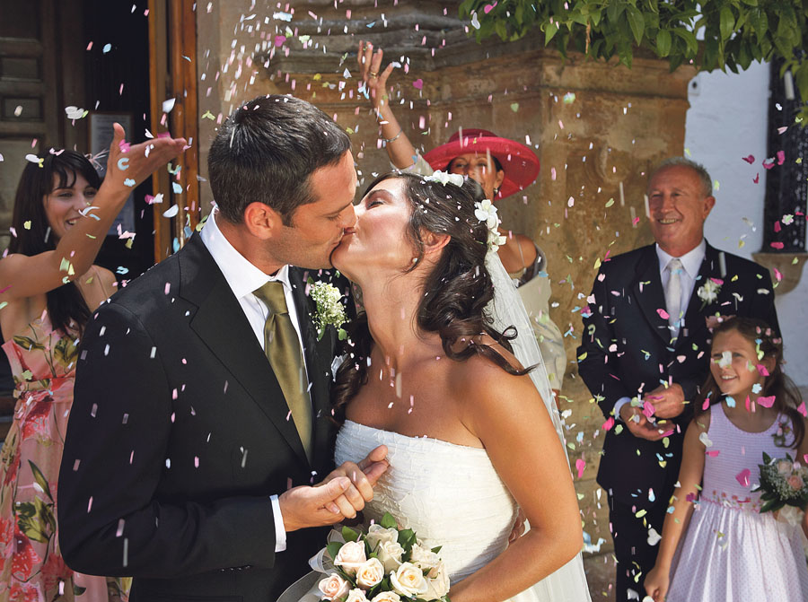 Guests throwing confetti over kissing bride and groom, outdoors