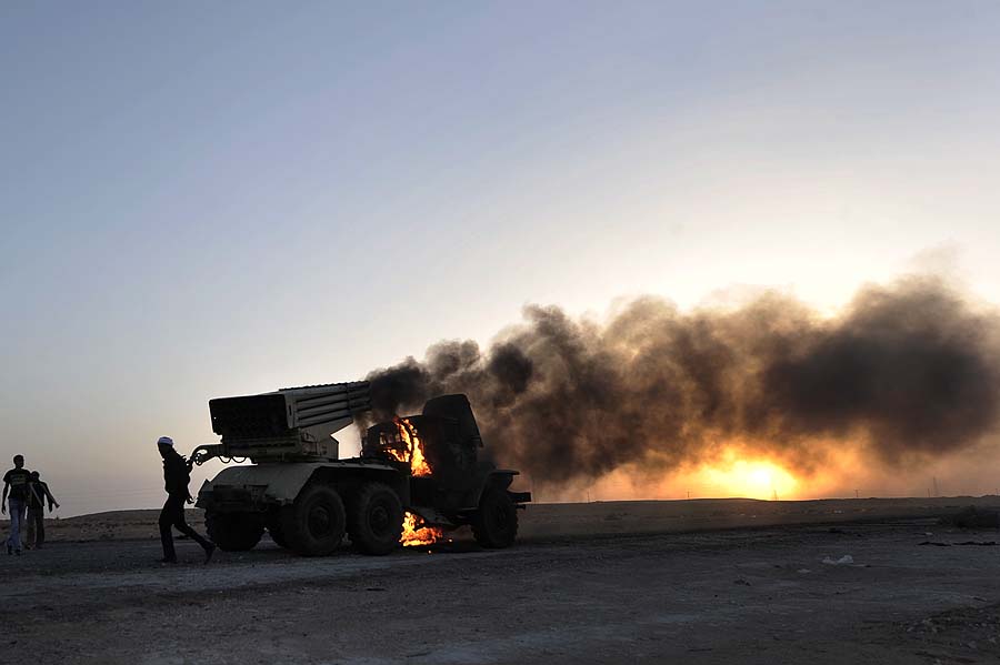 Libyans walk around a burning multi-rocket launcher outside the oil rich town of Ras Lanuf on March 27 2011, as they and press on as far as Nofilia with Sirte, 360 kilometres (225 miles) east of Tripoli, the home town of Libyan leader Moamer Kadhafi in their sights. AFP PHOTO/ARIS MESSINIS
