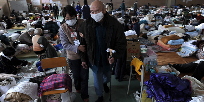An elderly Japanese tsunami survivor ia helped by a volonter as he walks through a gymnasium transformed as a temporary shelter at a relief centre in the tsunami-damaged city of Rikuzentakata, in Iwate prefecture on March 24, 2011. Shops and restaurants in sushi-mad Hong Kong, one of Japan's key food export markets, have been hit badly by the radiation scare with shoppers and diners deserting Japanese produce en masse.    AFP PHOTO/ NICOLAS ASFOURI