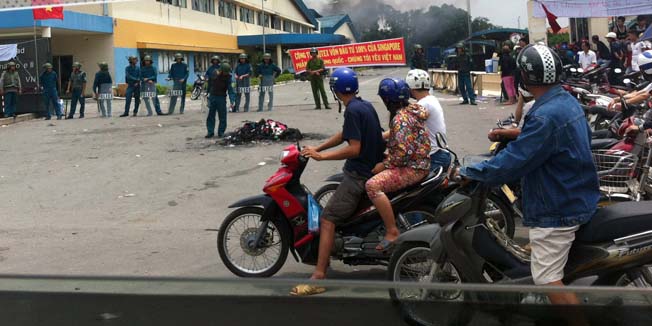 Commuters drive past as riot police (L) stand guard outside a burning factory building (back) in the Vietnamese southern province of Binh Duong on May 14, 2014, as anti-China protesters set more than a dozen factories on fire in Vietnam in the biggest eruption of rage against Beijing for decades over the deployment of an oil rig in contested waters. China expressed 