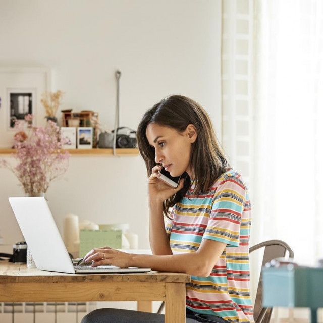 Beautiful woman talking through smart phone while using laptop at table. Mid adult female is using wireless technology while sitting in domestic kitchen. She is wearing casuals at home.