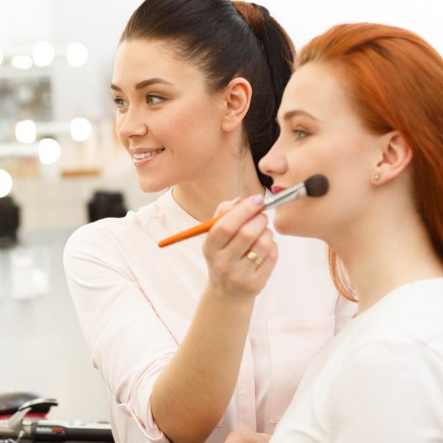 Cheerful young female makeup artist smiling enjoying working with a beautiful young red haired female model, applying makeup on her face using powder brush copy space