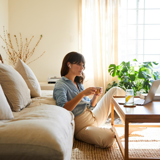 Young woman sitting on her living room floor at home wearing wireless earbuds and streaming something on a laptop