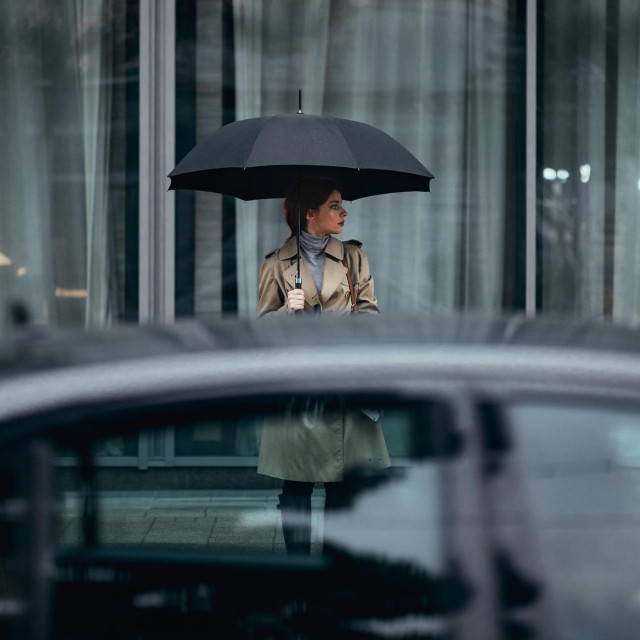 Young Caucasian woman with umbrella wearing trench coat and standing on sidewalk.