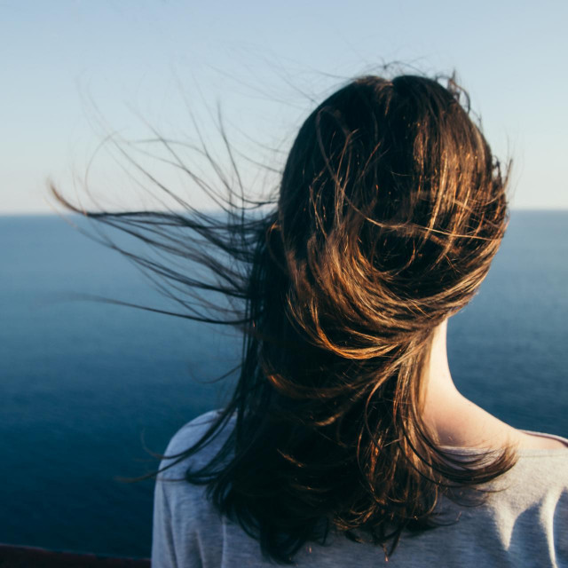 Woman with dark hair stands on a top cliff over blue sea view while wind.
