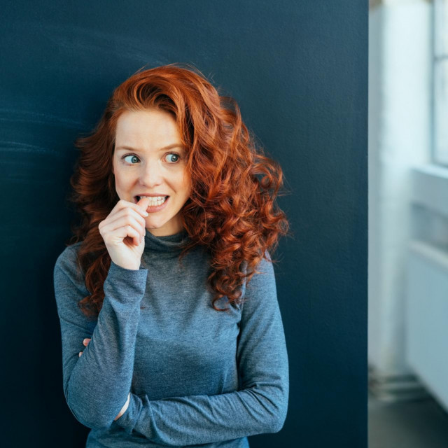 Worried attractive young woman with long wavy red hair standing indoors biting her nail in trepidation looking to the side