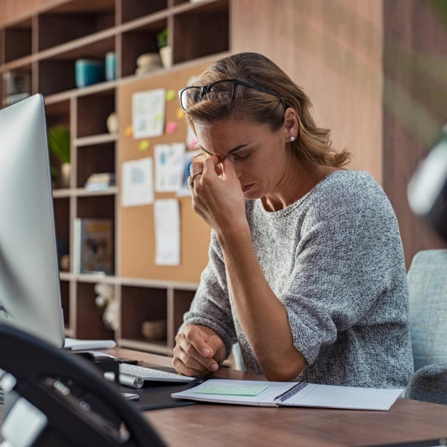 Exhausted businesswoman having a headache in modern office. Mature creative woman working at office desk with spectacles on head feeling tired. Stressed casual business woman feeling eye pain while overworking on desktop computer.
