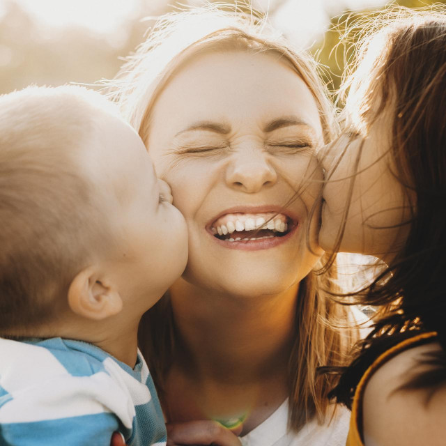 Close up portrait of lovely young mother laughing with closed eyes while her kids is kissing her on the cheeks outdoor against sunset.
