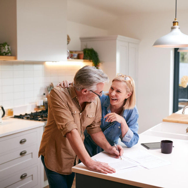 Mature Couple Reviewing And Signing Domestic Finances And Investment Paperwork In Kitchen At Home