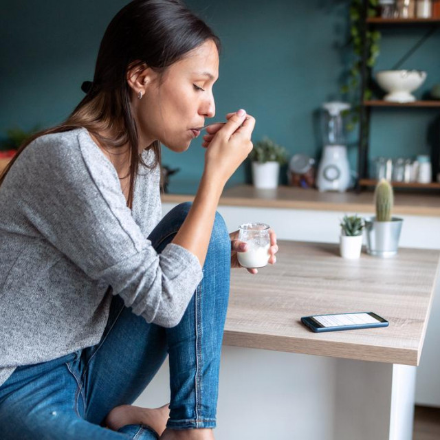 Shot of pretty young woman eating yogurt while using her mobile phone in the kitchen at home.
