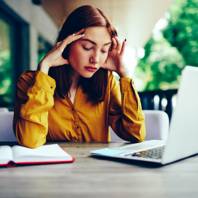 Beautiful skilled female graphic designer deeply thinking while distantly working on laptop computer connected to 4G.Tired redhead woman making remote job while sitting at wooden table outdoors