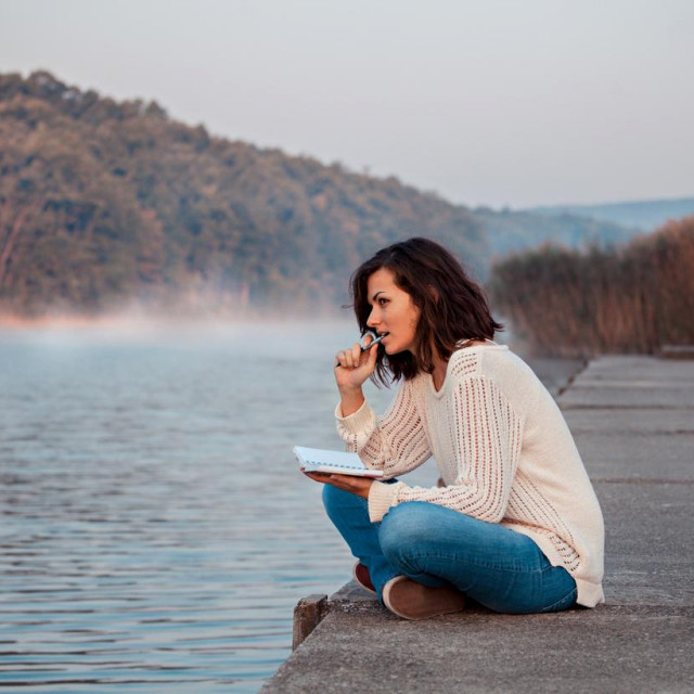 Thoughtful girl sitting by lake and writes diary