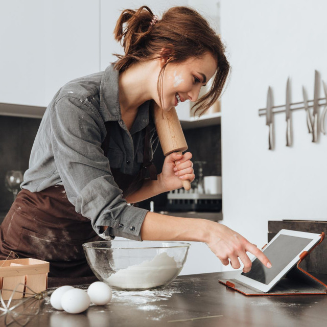 Image of young pretty lady standing in kitchen and cooking the dough. Looking at tablet computer.