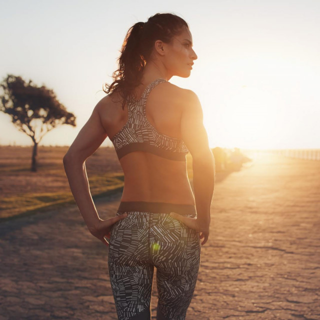 Rear view shot of fit woman in sportswear walking along a road and looking away. She is walking on seaside promenade during sunset ready to do fitness workout.