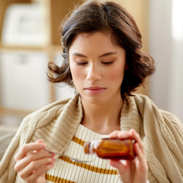 people, health and medicine concept - sad sick young woman in blanket pouring antipyretic or cough syrup from bottle to spoon at home
