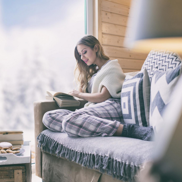 Young beautiful blonde woman with cup of coffee sitting home in living room by the window reading book. Winter snow landscape view. Christmas concept
