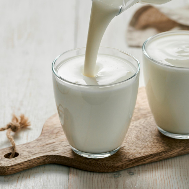 Pouring kefir, buttermilk or yogurt with probiotics. Yogurt flowing from glass bottle on white wooden background. Probiotic cold fermented dairy milk drink. Vertical
