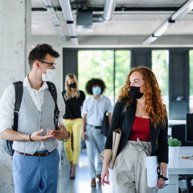 Young people with face masks back at work in office after lockdown, talking.
