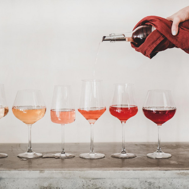 Shades of Rose wine in stemmed glasses placed in line from light to deep and womans hand pouring wine from bottle to glass, white wall background behind. Wine bar, wine shop, tasting concept