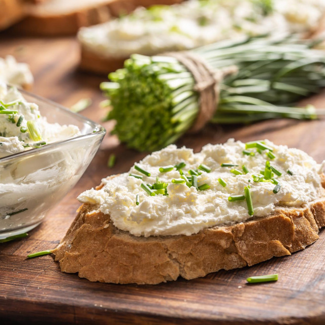 A bowl of homemade cream cheese spread with chopped chives surrounded by bread slices with spread and a bunch of freshly cut chives.