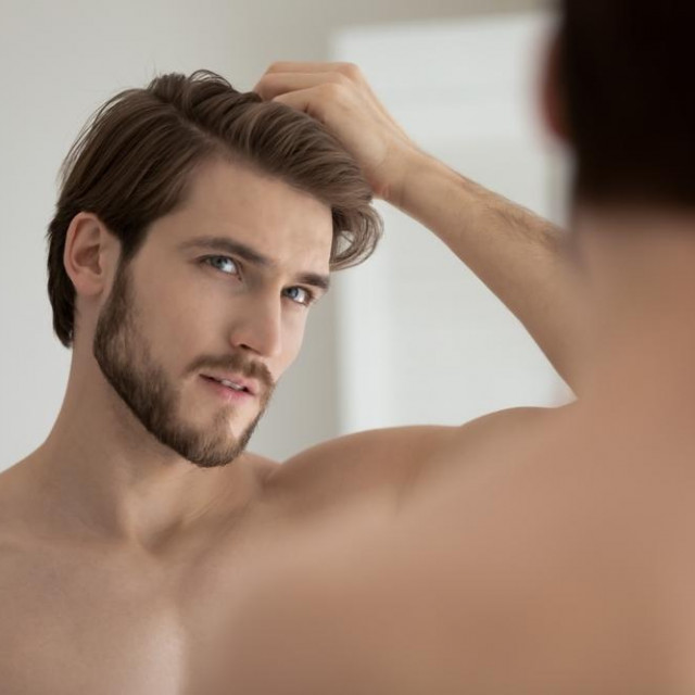 Young shirtless guy looks in mirror touch hair feels concerned due to receding, dry, dull hair. Hair loss common hair problem prevention, alopecia treatment ad, cosmetics products for male concept