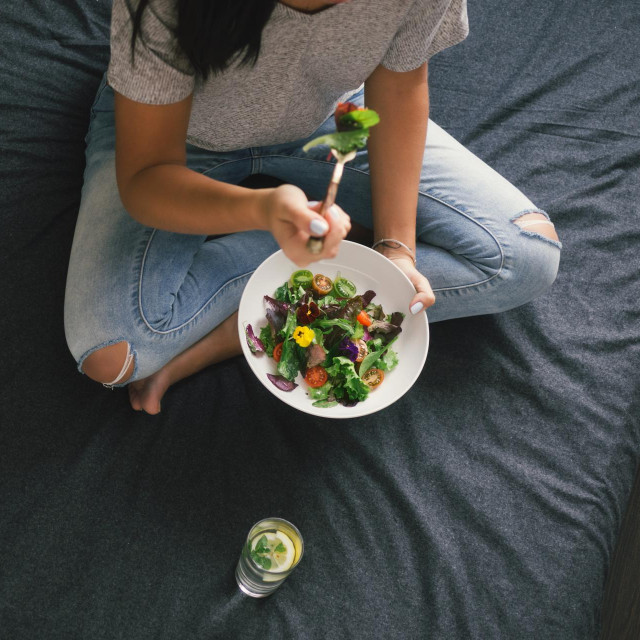 Young woman eating salad sitting on bed at home, top view