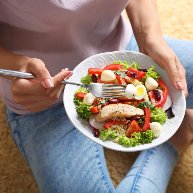 Young woman eating tasty chicken salad with vegetables while sitting on floor