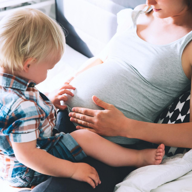 Pregnant mother and son are talking and spending time together at home. Little child boy looking at her mother pregnant tummy. Pregnancy, family, parenthood, preparation and expectation concepts.