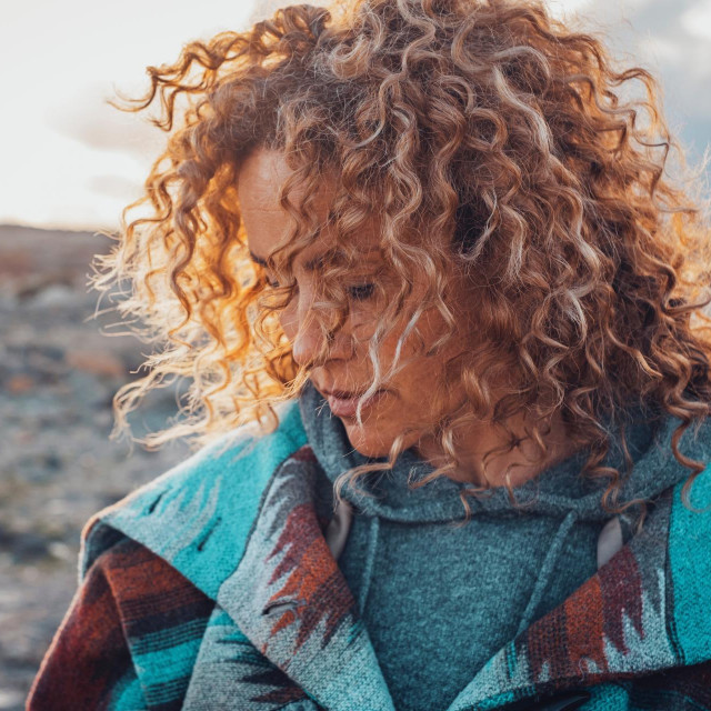 Side portrait of attractive woman with blonde coloured long curly hair looking down and enjoying outdoors nature feeling. Travel adult female people concept lifestyle with desert in background