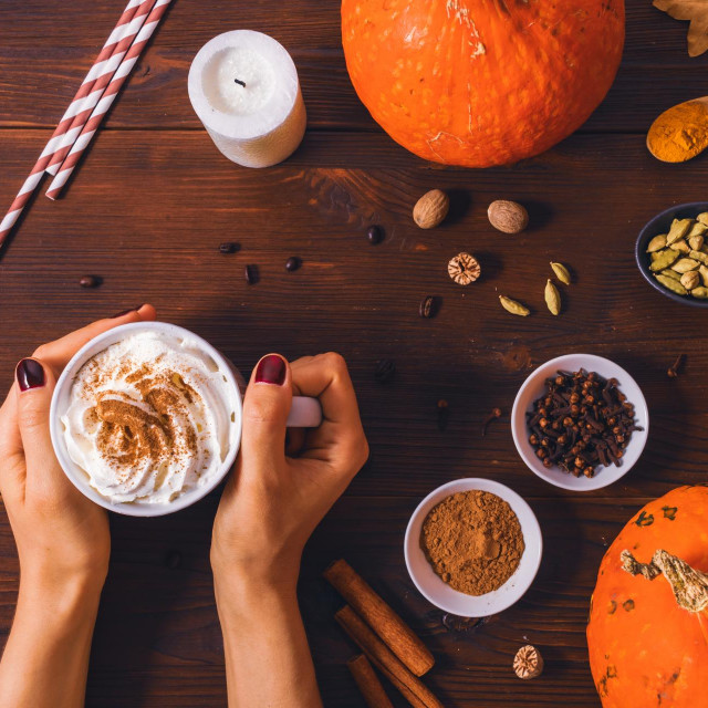 Autumn warm drink pumpkin spice latte in female hands next to organic seasoning ingredients on wooden table, top view