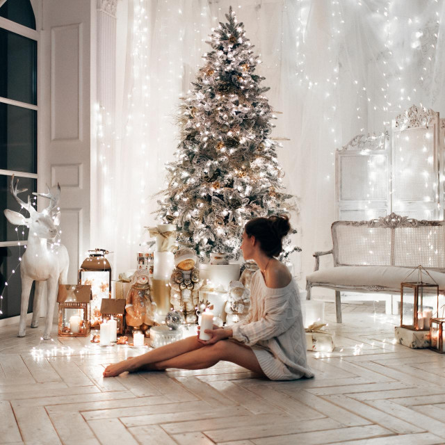 woman in white sweater, warm and cozy evening in Christmas interior design, Xmas tree decorated by lights gifts toys, candles, lanterns, garland lighting indoors fireplace.holiday living room.New year