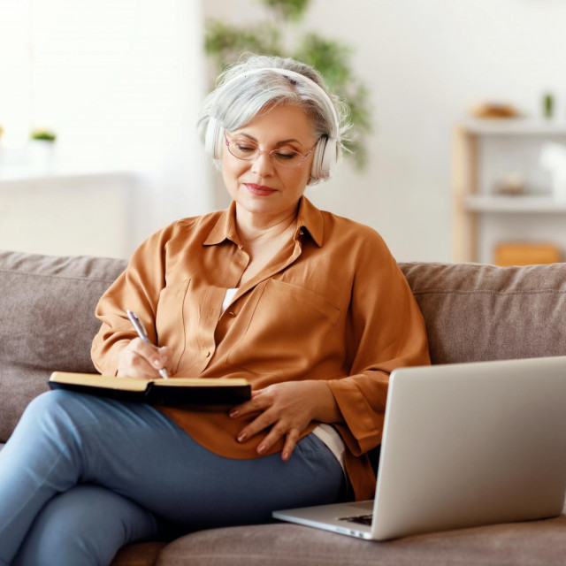 Elderly female in headphones and glasses writing in notebook and listening to music while sitting on couch near laptop