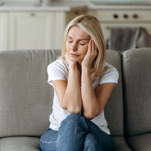 Headache, depressed or stress. Middle aged blonde woman sits on sofa at living room holding her head with her hands, feels unhappy because of headache, stress, depressed or bad news, she need rest and