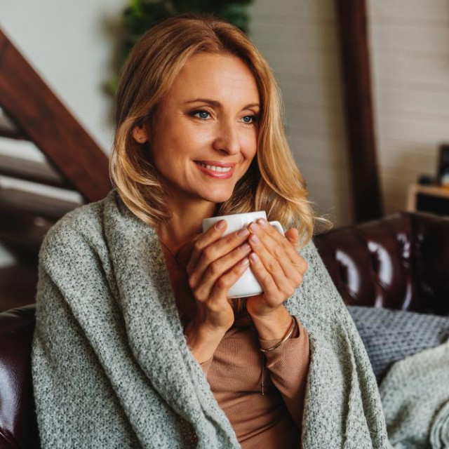 Close up of dreaming middle aged woman sitting in living room with cup of coffee or tea enjoying under blanket