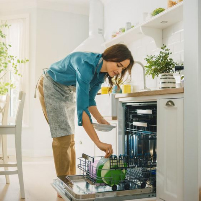 Beautiful Female is Loading Dirty Plates into a Dishwasher Machine in a Bright Sunny Kitchen. Girl in Wearing an Apron. Young Housewife Uses Modern Appliance to Keep the Home Clean.