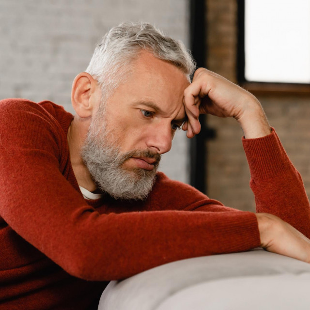 Sad stressed troubled mature middle-aged man sitting on the couch, missing his friends, feeling bad unwell unhealthy, suffering from depression at home