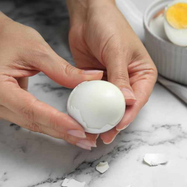 Woman peeling boiled egg at white marble table, closeup