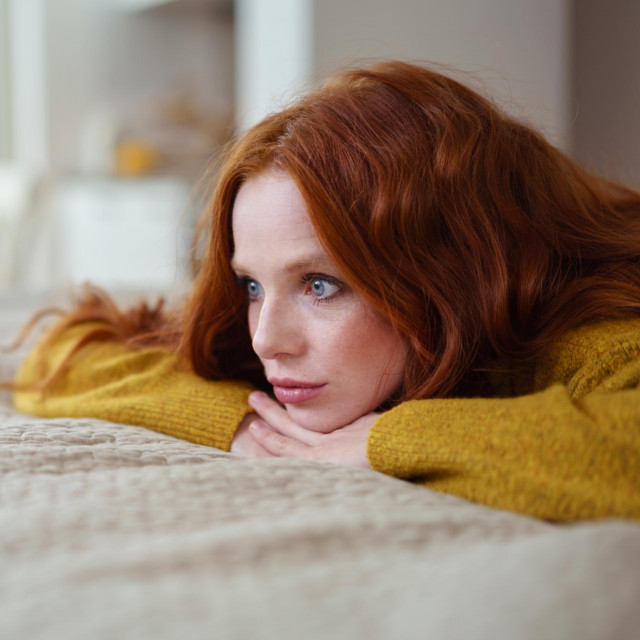 Attractive young redhead woman lying on her bed on her stomach thinking resting her chin on her hands and staring into the distance
