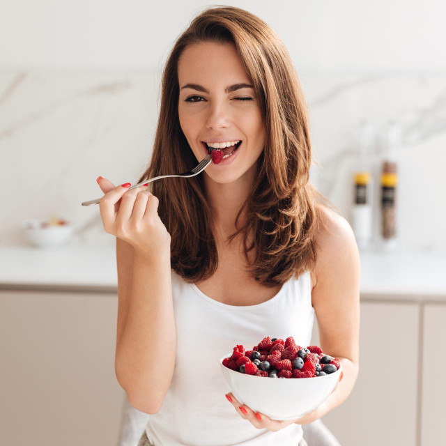 Playful young woman holding bowl with fresh berries while sitting in a kitchen and winking