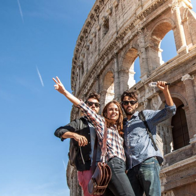 Three young friends tourists posing for funny pictures in front of colosseum in rome. Blue sky and lens flare on sunny day.