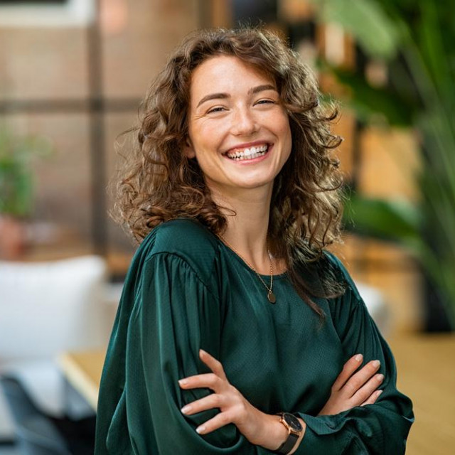 Portrait of young smiling woman looking at camera with crossed arms. Happy girl standing in creative office. Successful businesswoman standing in office with copy space.