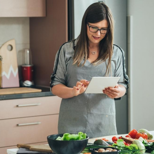Happy woman using digital tablet for watching internet tutorials and making healthy meal while standing in front of the table full with fresh organic vegetables in the kitchen