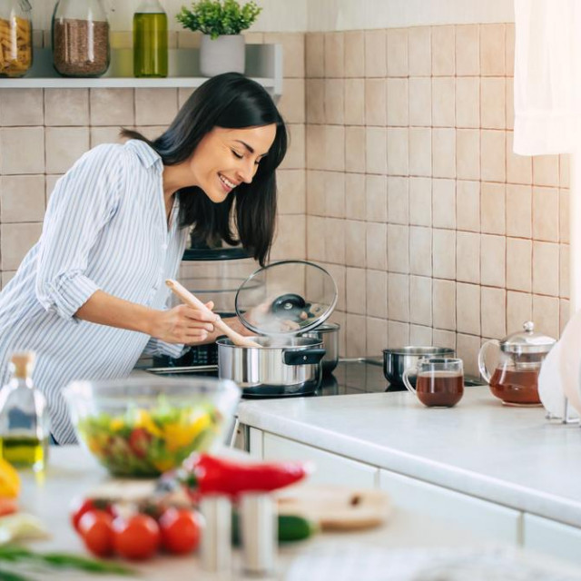 Beautiful happy young woman is cooking in the home kitchen and testing some soup from the pan on the stove
