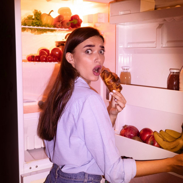 Failure of diet. Young woman eating junk croissant near opened fridge late in night, free space