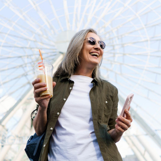 Cheerful senior woman enjoys a sunny day in an old European city, holding her phone and a cup of juice in her hands with a big smile on her face, embodying the concept of elderly happiness. High