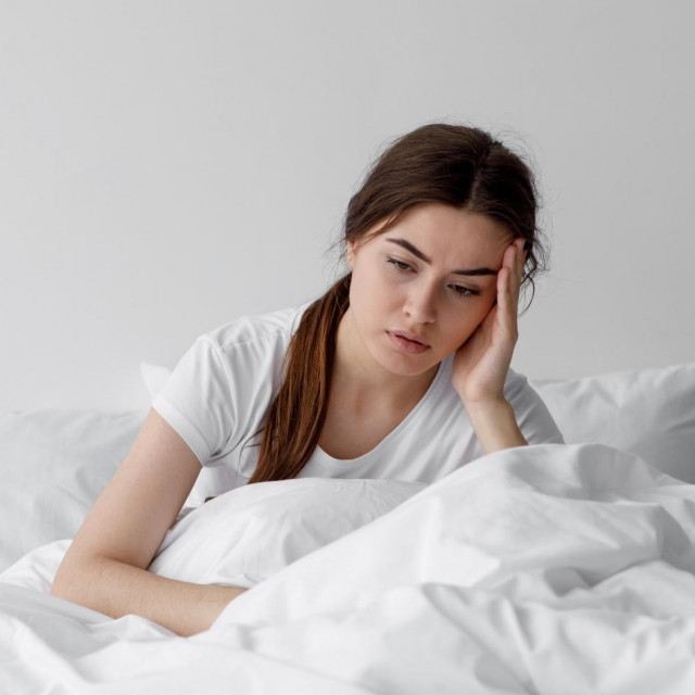 Sad european millennial female, worried, suffers from insomnia and headache. Lady sits on bed in bedroom at home, copy space. Migraines, health and mental problems, lack of sleep and depression
