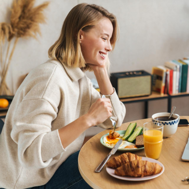 Photo of happy blonde woman smiling and using laptop while having breakfast in cozy living room