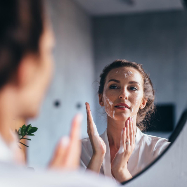 Woman washes in front of the mirror, applying foam to her face.