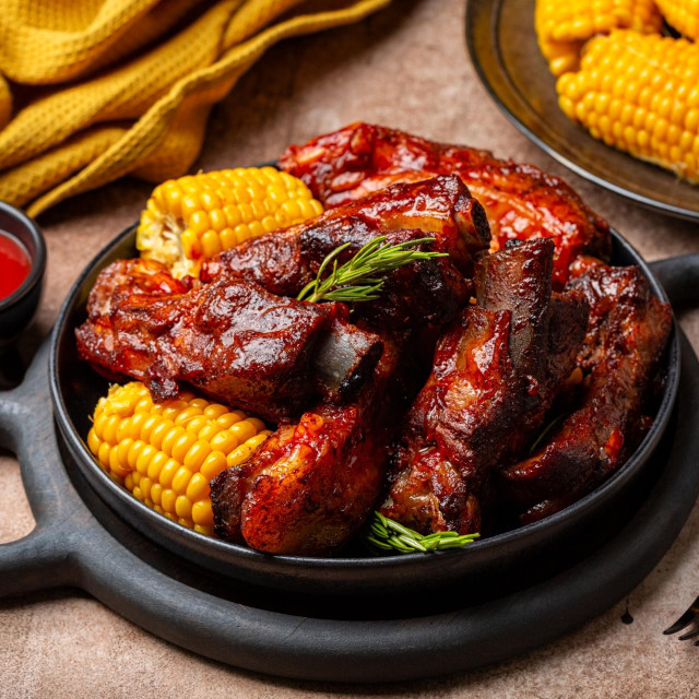 Food board with baked pork ribs with corn cobs, barbecue sauce, rosemary. Brown table background. Meat dinner.
